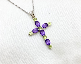 Puede incluir: Un collar de cadena de plata con un colgante de cruz. La cruz está adornada con piedras preciosas ovaladas de color morado y verde.