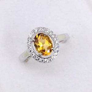 Peut inclure: Une bague en argent avec une grande pierre précieuse jaune de forme ovale entourée d'un halo de petites pierres précieuses blanches.