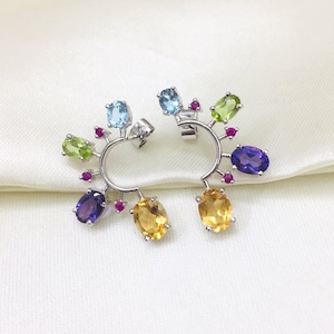 Puede incluir: Un par de pendientes de aro de plata con una variedad de piedras preciosas de colores. Los pendientes presentan una mezcla de piedras preciosas azules, verdes, amarillas y moradas.