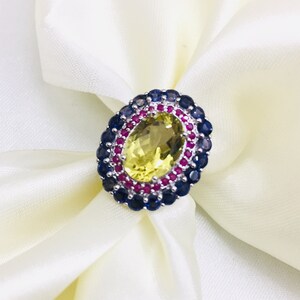 May include: A silver ring with a large oval-shaped yellow gemstone surrounded by a double row of smaller red and blue gemstones.