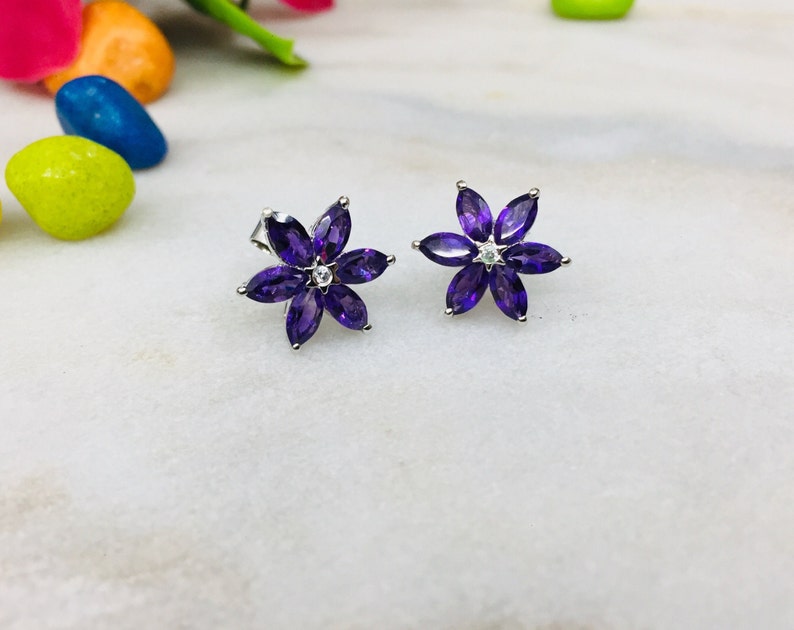 May include: A pair of silver flower-shaped earrings with purple gemstones and a small clear gemstone in the center of each flower.