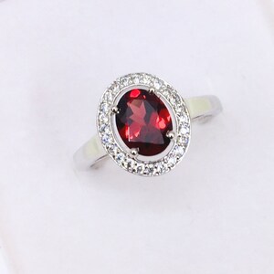 Peut inclure: Une bague en argent avec une grande pierre ovale rouge entourée d'un halo de petites pierres blanches.