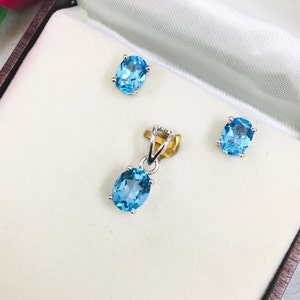 May include: A jewelry set featuring a pendant and matching stud earrings. The pendant and earrings each have a light blue, oval-shaped gemstone set in a silver-colored metal. The set is displayed in a white satin-lined box.