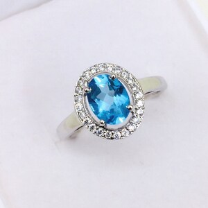 Peut inclure: Une bague en argent ornée d'une grande pierre précieuse ovale bleu vif, entourée d'un halo de petites pierres précieuses claires. La bague a un anneau simple en argent poli. La bague est posée sur un fond blanc.