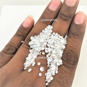 Op de afbeelding: Een hand die een grote stapel kleine, heldere, ronde diamanten vasthoudt. De diamanten zijn verspreid over de hand en vallen op het oppervlak eronder. De tekst "@AMBAJEWEL" is zichtbaar in de afbeelding.