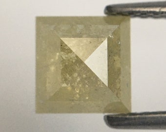 Puede incluir: Un diamante cuadrado de color amarillo claro con un corte en forma de pirámide. El diamante tiene una superficie rugosa y texturizada.