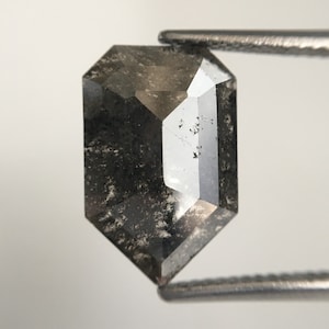 Könnte beinhalten: Ein facettierter, rechteckiger Diamant mit Salz-und-Pfeffer-Optik. Die Edelstein wird von einer Pinzette gehalten, die seine einzigartigen inneren Einschlüsse und reflektierenden Oberflächen zeigt. Die Farbe des Diamanten ist eine Mischung aus Grau und Schwarz.