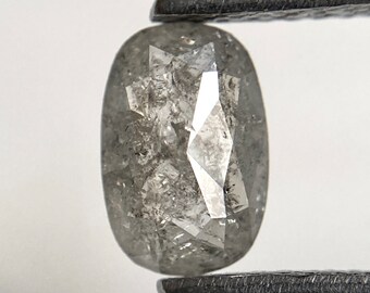 May include: A gray, oval-shaped diamond held by a pair of tweezers. The diamond has a faceted surface with internal inclusions, creating a unique, textured appearance. The gem is a neutral color, suitable for various jewelry designs.