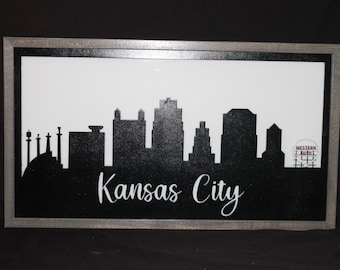 May include: Black and white silhouette of the Kansas City skyline with the text "Kansas City" below. The silhouette includes the Country Club Plaza, the Power & Light District, and the Sprint Centre. The silhouette is framed in a grey wood frame.