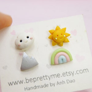 May include: A set of three colorful clay earrings featuring a smiling cloud, a yellow sun, and a rainbow. The earrings are handmade and are displayed on a white card with the text "www.beprettyme.etsy.com Handmade by Anh Dao."