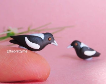 May include: Two small black and white birds with orange eyes made of polymer clay. The birds are perched on a finger and a pink surface. The birds are a popular Australian species.