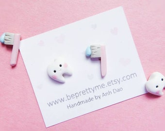 May include: A pair of handmade polymer clay earrings featuring a white tooth with a smiling face and a pink toothbrush with blue bristles. The earrings are displayed on a white card with the text "www.beprettyme.etsy.com Handmade by Anh Dao".
