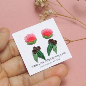 May include: A pair of handmade polymer clay earrings featuring pink flowers and green leaves with brown gum nuts. The earrings are displayed on a white card with the text "www.beprettyme.etsy.com Handmade by Anh Dao".