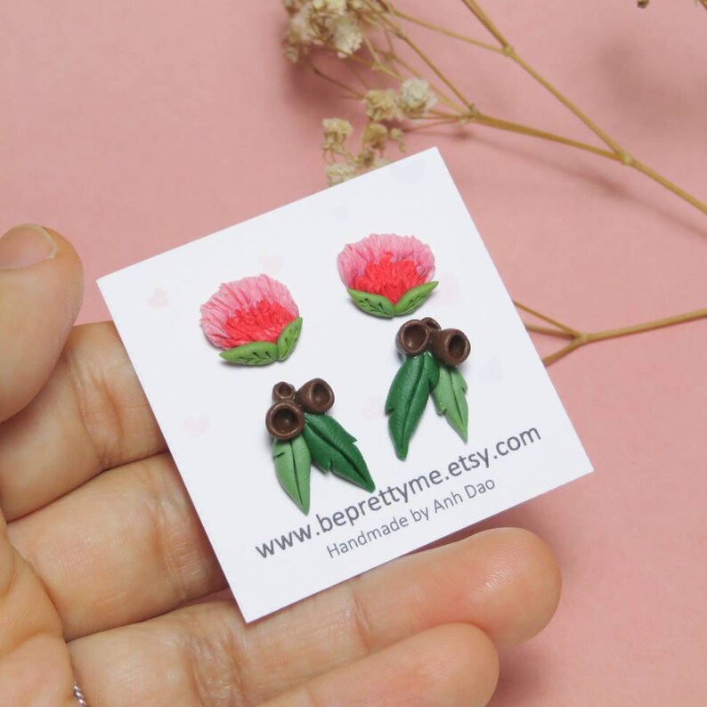 May include: A pair of handmade polymer clay earrings featuring pink flowers and green leaves with brown gum nuts. The earrings are displayed on a white card with the text "www.beprettyme.etsy.com Handmade by Anh Dao".