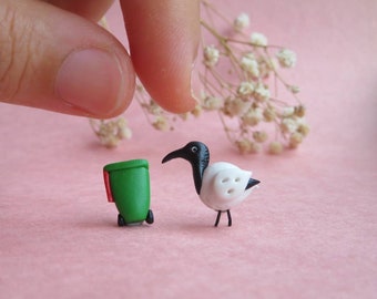 May include: A small, white bird with a black head and beak stands next to a miniature green trash can. The bird has a button-like detail on its chest. The scene is set against a pink background.