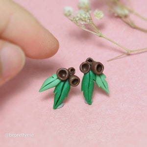 May include: A pair of earrings featuring a cluster of brown gum nuts with green leaves. The earrings are made of polymer clay and are a unique and stylish accessory.