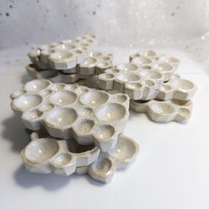 May include: A collection of off-white ceramic paint palettes. Each palette features a honeycomb pattern with numerous circular wells of different sizes. The palettes have a speckled texture and a glossy finish, indicating they are handmade.