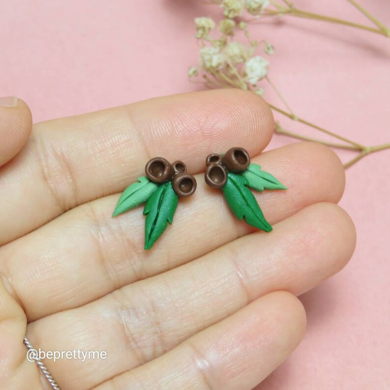 May include: A pair of earrings featuring small brown and green clay leaves with brown clay acorns. The earrings are on a hand with a pink background.