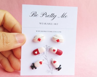 May include: A set of three pairs of earrings featuring a nurse's hat, a pill, and a syringe. The earrings are made of white and red polymer clay and have a black accent. The earrings are displayed on a white card with the text "Be Pretty Me Wearable Art www.beprettyme.etsy.com".