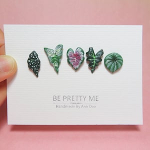 May include: Six small, handmade polymer clay earrings shaped like different types of leaves. The leaves are green, white, and pink. The earrings are displayed on a white card with the text "BE PRETTY ME Handmade by Anh Dao".