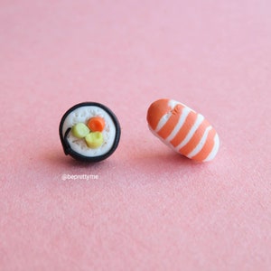 Könnte beinhalten: Ein Paar Sushi-Ohrringe. Ein Ohrring ist eine schwarz-weiße Maki-Rolle mit orangefarbener und grüner Füllung. Der andere Ohrring ist ein Lachs-Nigiri mit orange-weißen Streifen.
