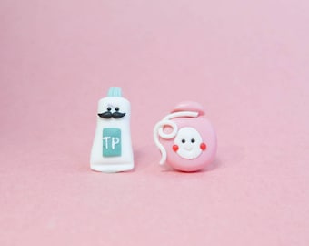 May include: A pair of miniature clay earrings, one shaped like a tube of toothpaste with a mustache and the letters "TP" on it, and the other shaped like a pink bottle of mouthwash with a smiling face.