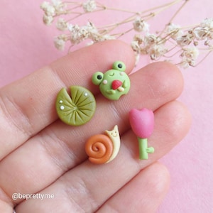 May include: A set of four miniature clay charms featuring a green frog holding a red mushroom, a green lily pad, a brown snail with a yellow shell, and a pink tulip.