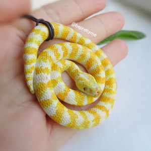 May include: A yellow and white snake charm necklace. The snake is coiled in a circle and has a black cord.