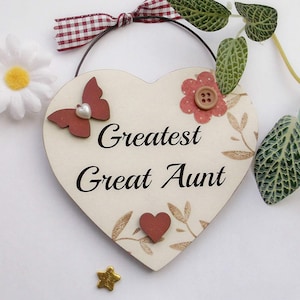 Può includere: Un cartello di legno bianco a forma di cuore con il testo "Greatest Great Aunt" in lettere marroni. Il cartello è decorato con una farfalla rossa, un fiore rosso con un bottone marrone e un cuore marrone. Il cartello è appeso a uno spago marrone.