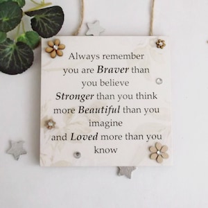 May include: White wooden sign with a motivational quote: "Always remember you are Braver than you believe Stronger than you think more Beautiful than you imagine and Loved more than you know". The sign is decorated with small wooden flowers and stars.