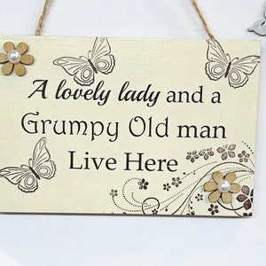 May include: A wooden sign with a cream background and brown text that reads "A lovely lady and a grumpy old man live here". The sign is decorated with brown butterflies and flowers.