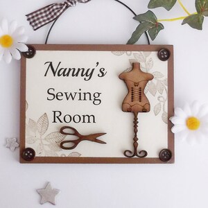 May include: A wooden sign with a brown frame and a white background. The sign says "Nanny's Sewing Room" and has a wooden cutout of a dress form and a pair of scissors.
