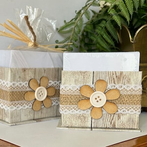 Puede incluir: Dos cajas de madera con un diseño de veta de madera rústica. Cada caja tiene un borde de encaje blanco y una gran flor de tela marrón con un botón blanco en el centro. Las cajas están vacías y listas para ser llenadas.