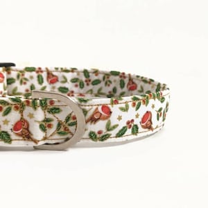 May include: White dog collar with a festive Christmas pattern. The fabric features red robins, green leaves, gold stars, and red berries. The collar has a black plastic buckle and a silver metal D-ring.