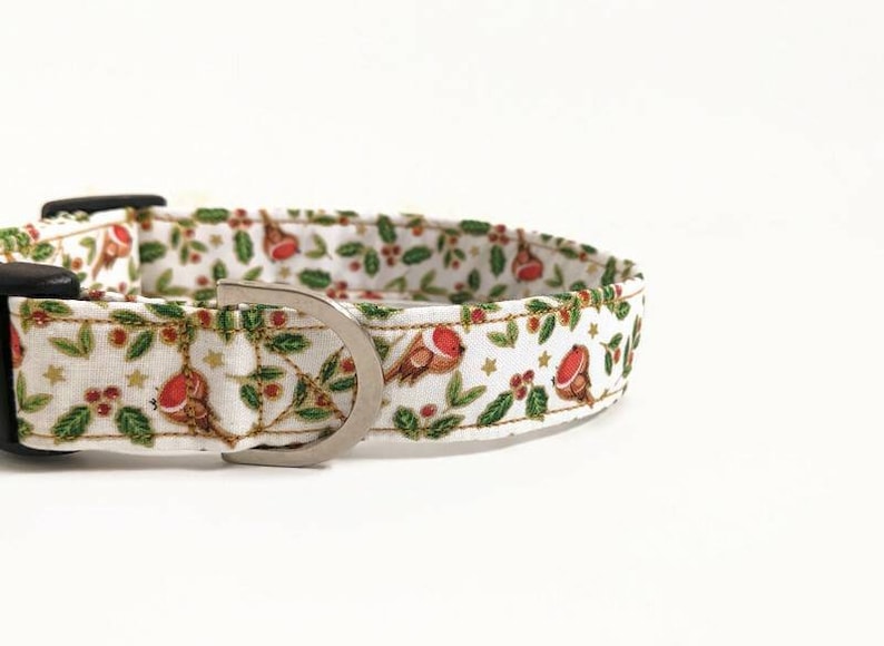 May include: White dog collar with a festive Christmas pattern. The fabric features red robins, green leaves, gold stars, and red berries. The collar has a black plastic buckle and a silver metal D-ring.