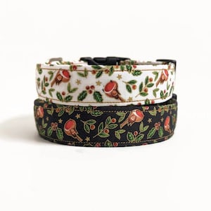 May include: Two dog collars, one stacked on top of the other. The top collar is white with a pattern of red birds, green leaves, and gold stars. The bottom collar is black with the same bird and leaf pattern. Both collars have black plastic buckles.