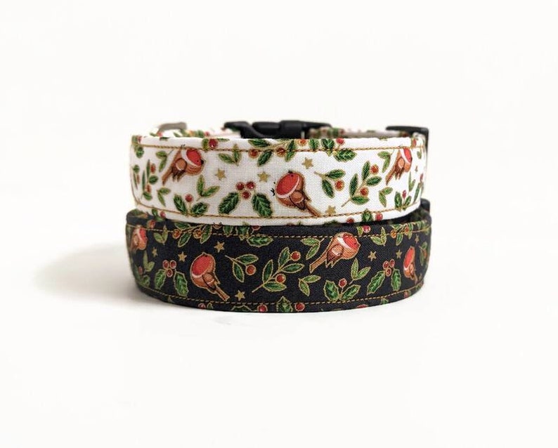 May include: Two dog collars, one stacked on top of the other. The top collar is white with a pattern of red birds, green leaves, and gold stars. The bottom collar is black with the same bird and leaf pattern. Both collars have black plastic buckles.