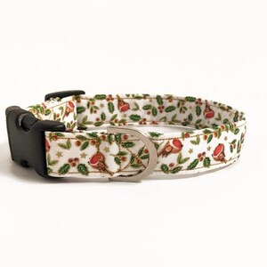 May include: A white dog collar with a festive pattern featuring red robins, green holly leaves, and gold stars. The collar has a black plastic buckle and a silver metal D-ring.
