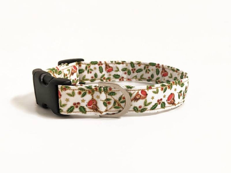 May include: A white dog collar with a festive pattern featuring red robins, green holly leaves, and gold stars. The collar has a black plastic buckle and a silver metal D-ring.