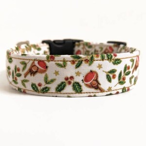 May include: A white dog collar with a festive pattern featuring brown robins with red breasts, green leaves, red berries, and gold stars. The collar has a black plastic buckle.