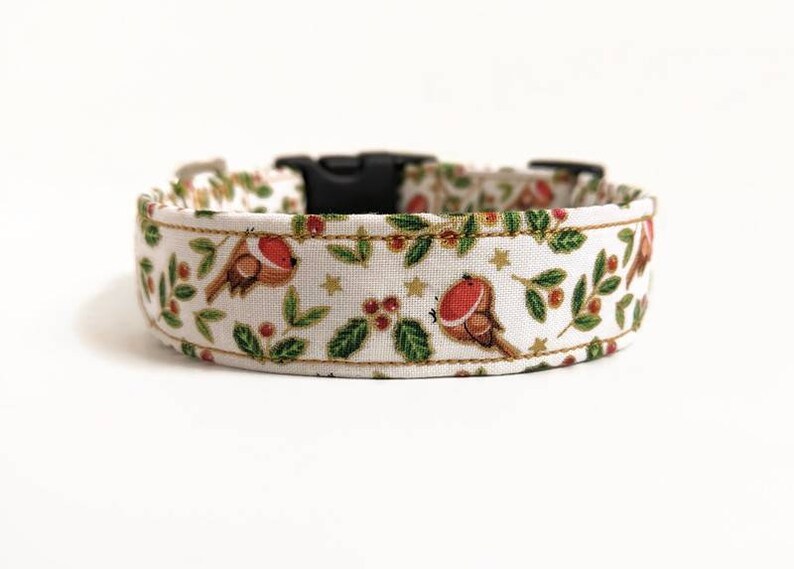 May include: A white dog collar with a festive pattern featuring brown robins with red breasts, green leaves, red berries, and gold stars. The collar has a black plastic buckle.
