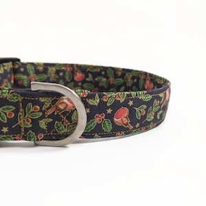 May include: A black dog collar with a festive pattern featuring red robins, green holly leaves, and gold stars. The collar has a silver metal D-ring and black plastic buckle. The fabric is a dark blue color.