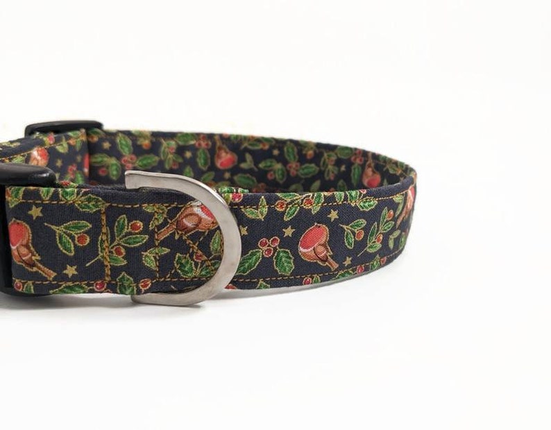 May include: A black dog collar with a festive pattern featuring red robins, green holly leaves, and gold stars. The collar has a silver metal D-ring and black plastic buckle. The fabric is a dark blue color.