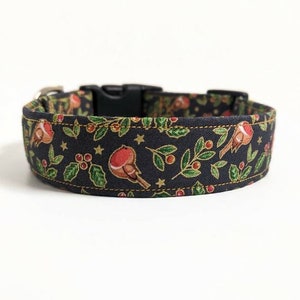 May include: A dark blue dog collar with a festive pattern featuring red birds, holly, berries, and gold stars. The collar has a black plastic buckle and a silver metal loop for leash attachment. The fabric is stitched with gold thread.