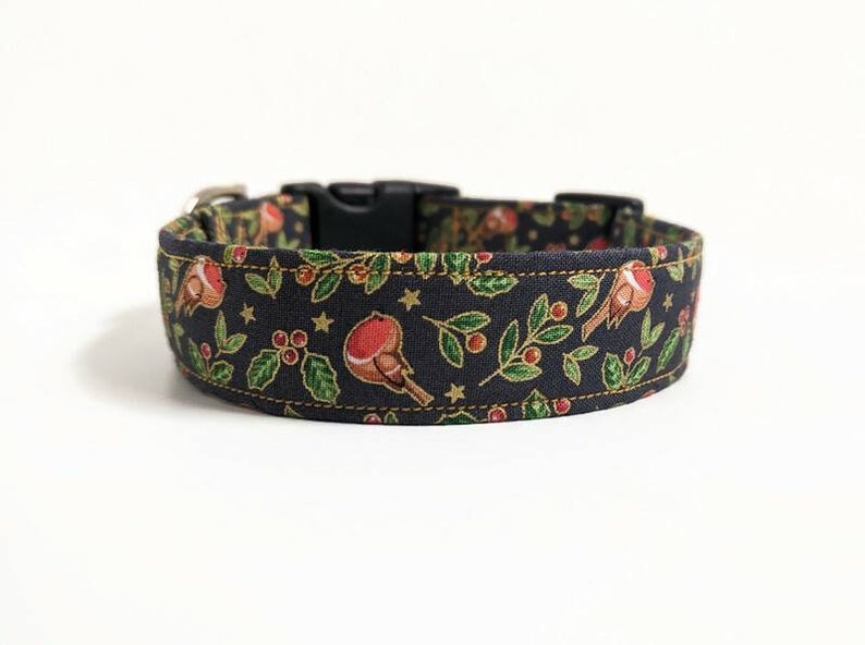 May include: A dark blue dog collar with a festive pattern featuring red birds, holly, berries, and gold stars. The collar has a black plastic buckle and a silver metal loop for leash attachment. The fabric is stitched with gold thread.