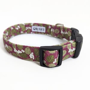 May include: A pink and green floral dog collar with a black buckle. The collar has a white background with green leaves and pink flowers.