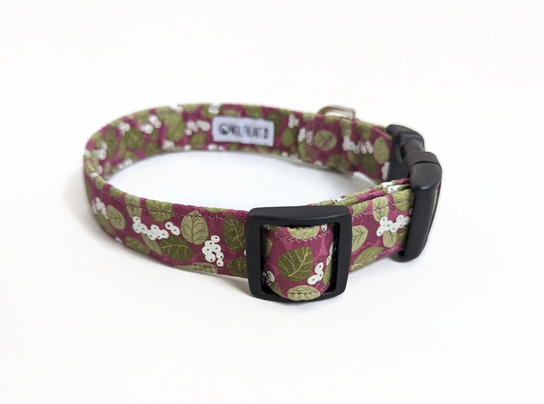 May include: A pink and green floral dog collar with a black buckle. The collar has a white background with green leaves and pink flowers.