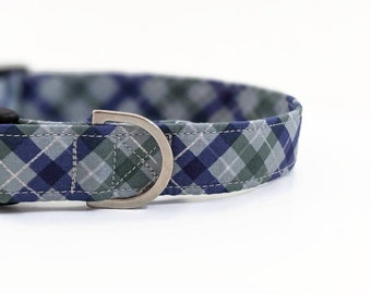 May include: Close-up of a dog collar featuring a blue, green, and grey plaid design. The collar has a silver metal D-ring and a black plastic buckle. The fabric is stitched with white thread.