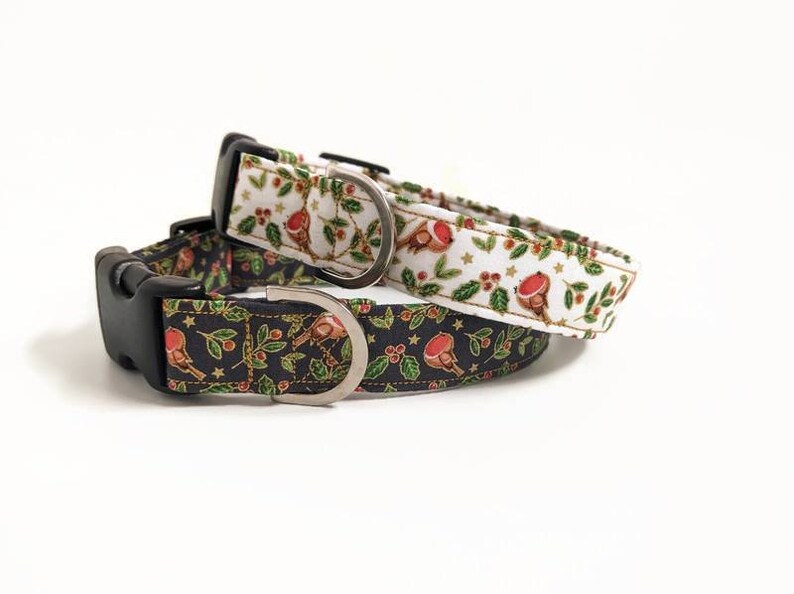 May include: Two Christmas-themed dog collars. One collar is white with red robins, green leaves, and red berries. The other is black with the same design. Both have black plastic buckles and silver metal D-rings.