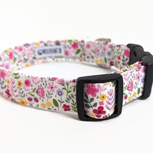 May include: A white dog collar with a floral print featuring pink, yellow, and blue flowers. The collar has a black plastic buckle.
